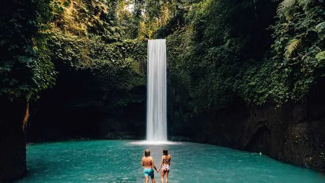 Visitors at the base of Tibumana Waterfall near Ubud Bali with tropical palm trees and lush green jungle surrounding the turquoise pool