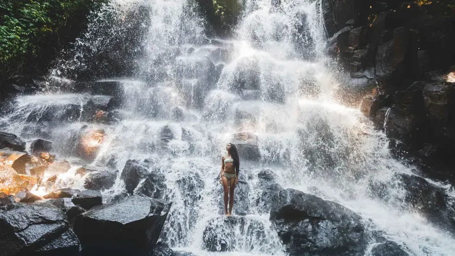Kanto Lampo Waterfall's tiered black rock face and shallow pool near Ubud Bali with a visitor posing on the rock ledges